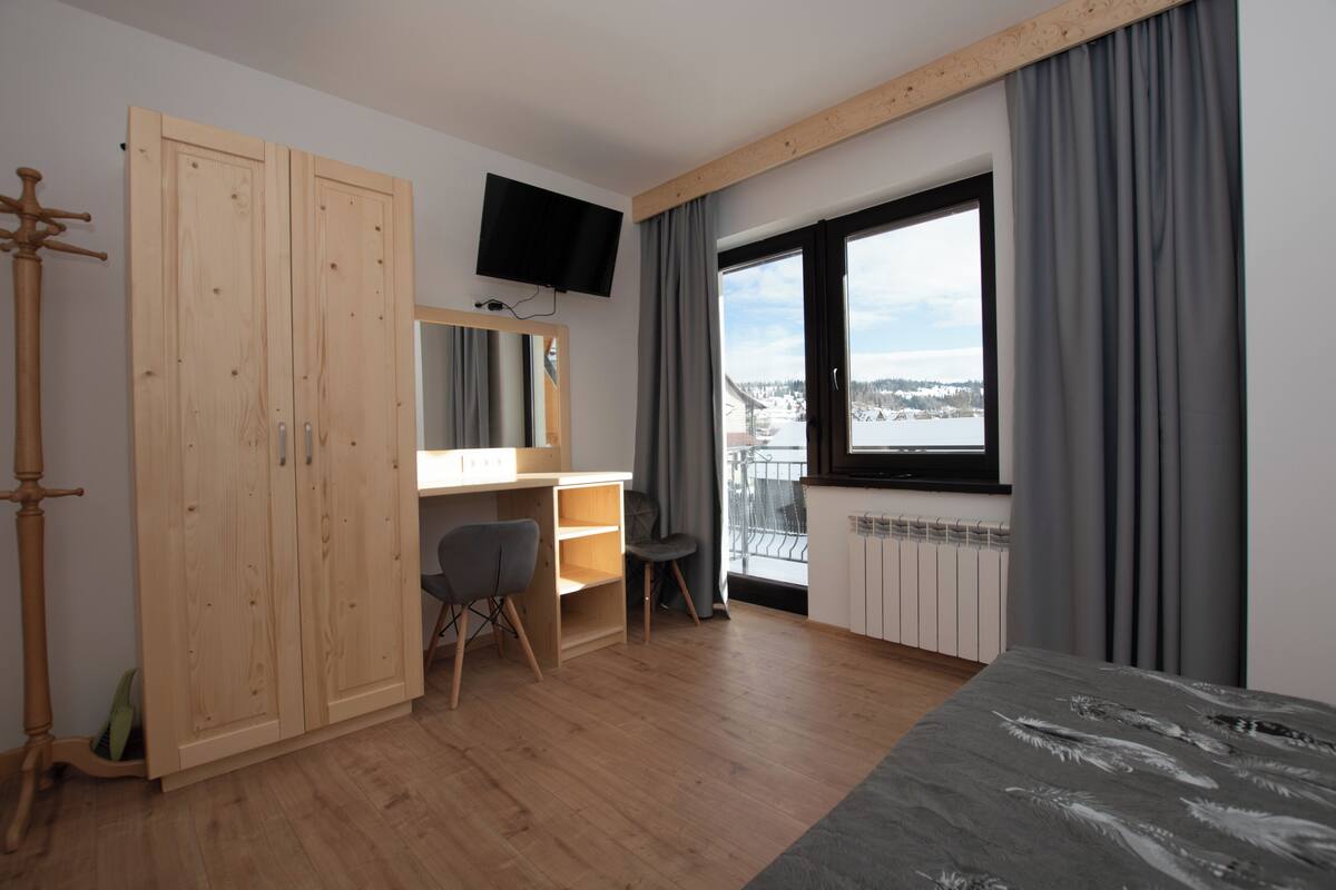 A bedroom with a wooden wardrobe, a desk with a chair, a television, a large window with curtains, and a view of a snowy landscape.