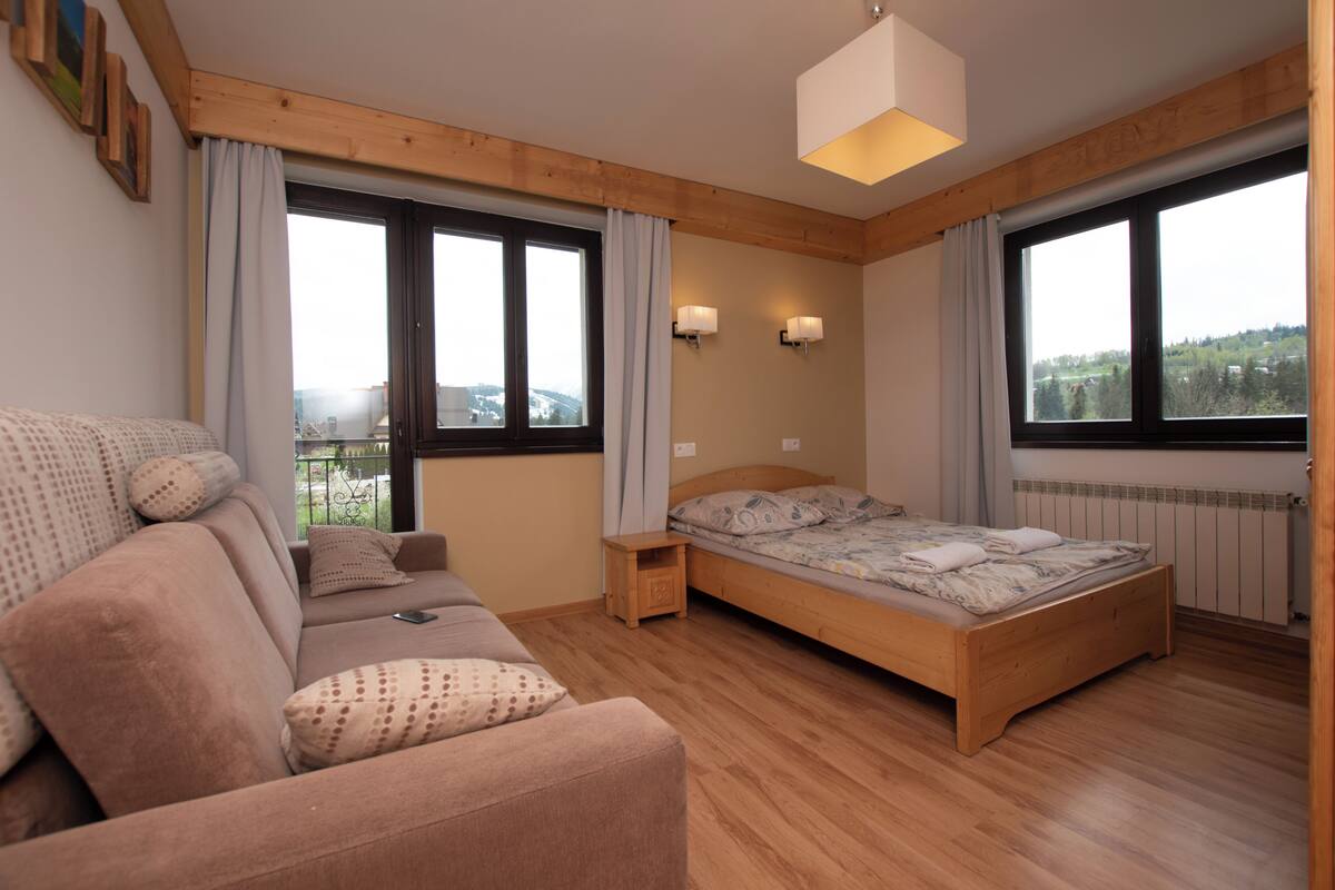 A bedroom with a wooden bed, a sofa, two bedside tables, and large windows offering a view of the outdoors.