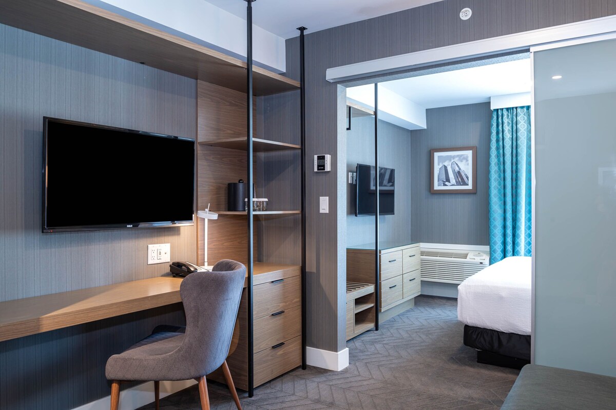 A modern hotel room with a flat-screen TV, a desk with a chair, a bed, a wardrobe, and a bathroom with a shower and toilet.
