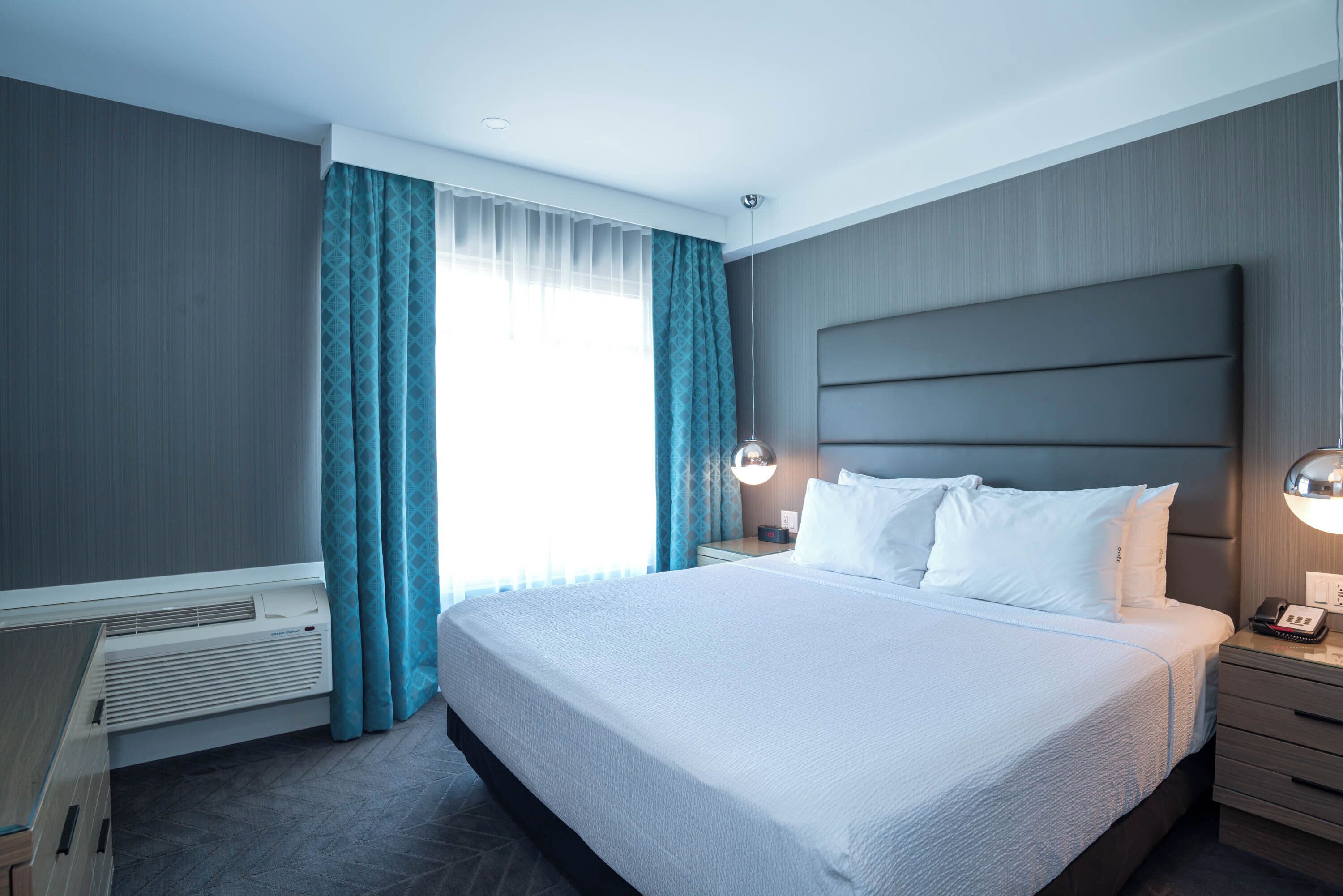 Habitación de hotel con una cama grande, una mesita de noche con lámpara, un calentador y una ventana con cortinas.