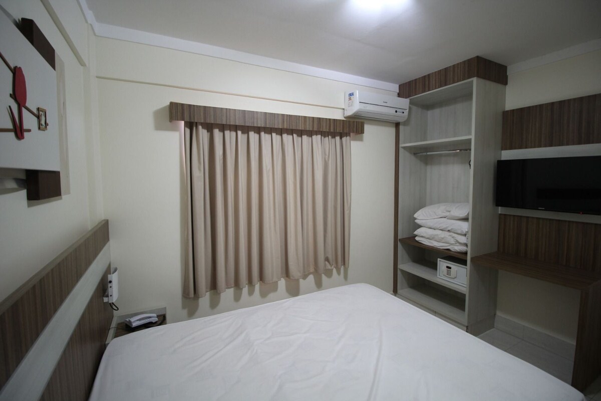 A hotel room with a bed, an air conditioning unit, a TV, and a wardrobe.