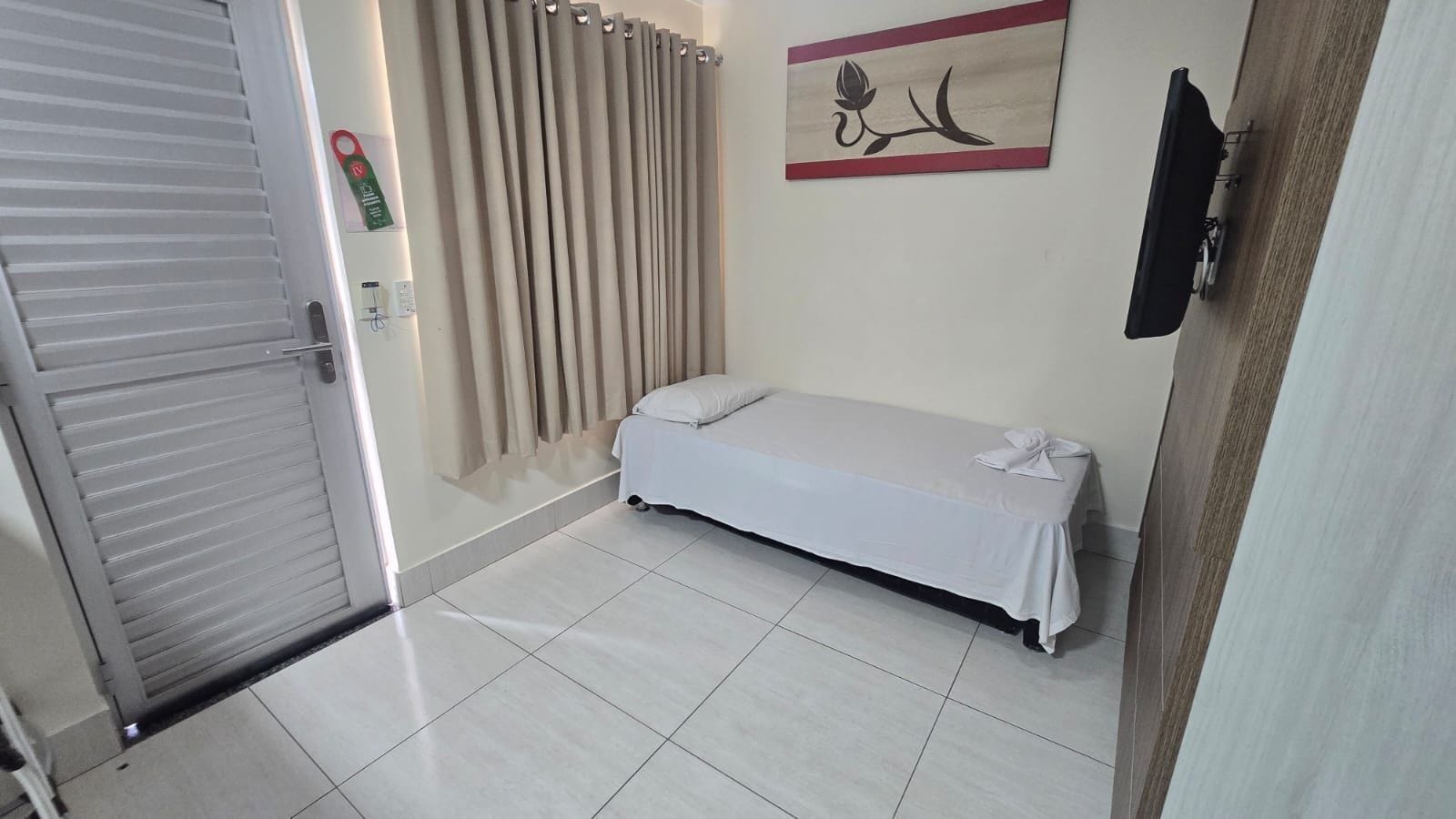 Quarto com cama, cortina, TV fixada na parede e porta.