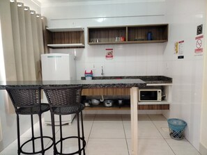 A kitchen with a granite countertop, two wicker chairs, a white refrigerator, a microwave, and a trash bin. - Apartment Lacqua DiRoma IV - JC Temporada (Caldas Novas)