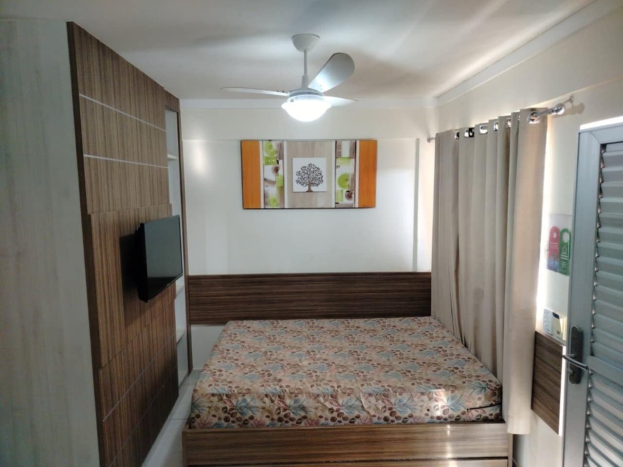 A bedroom with a bed, a ceiling fan, a wall-mounted TV, and a door with a curtain.