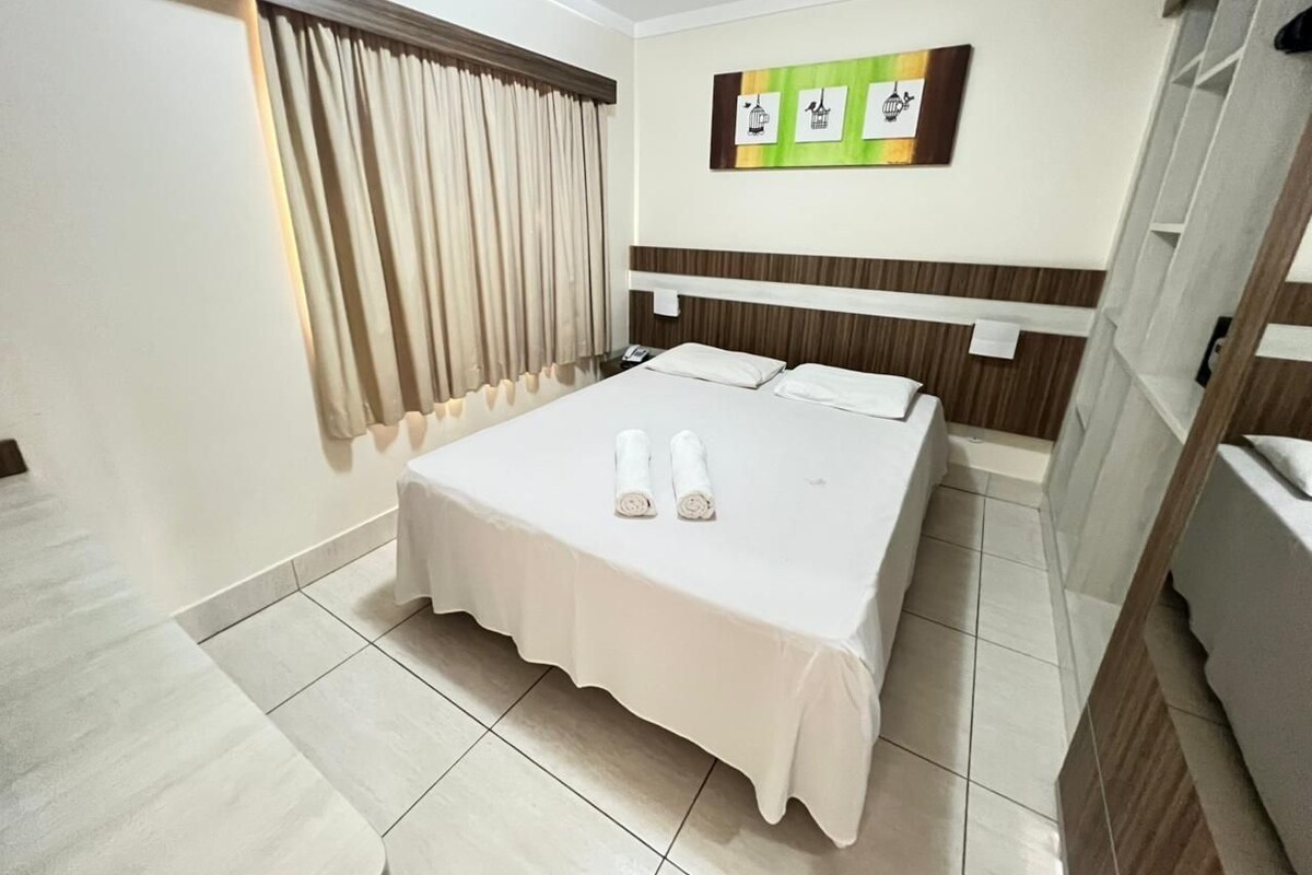 A hotel room with a bed, a window with curtains, a wall-mounted artwork, and a tiled floor.
