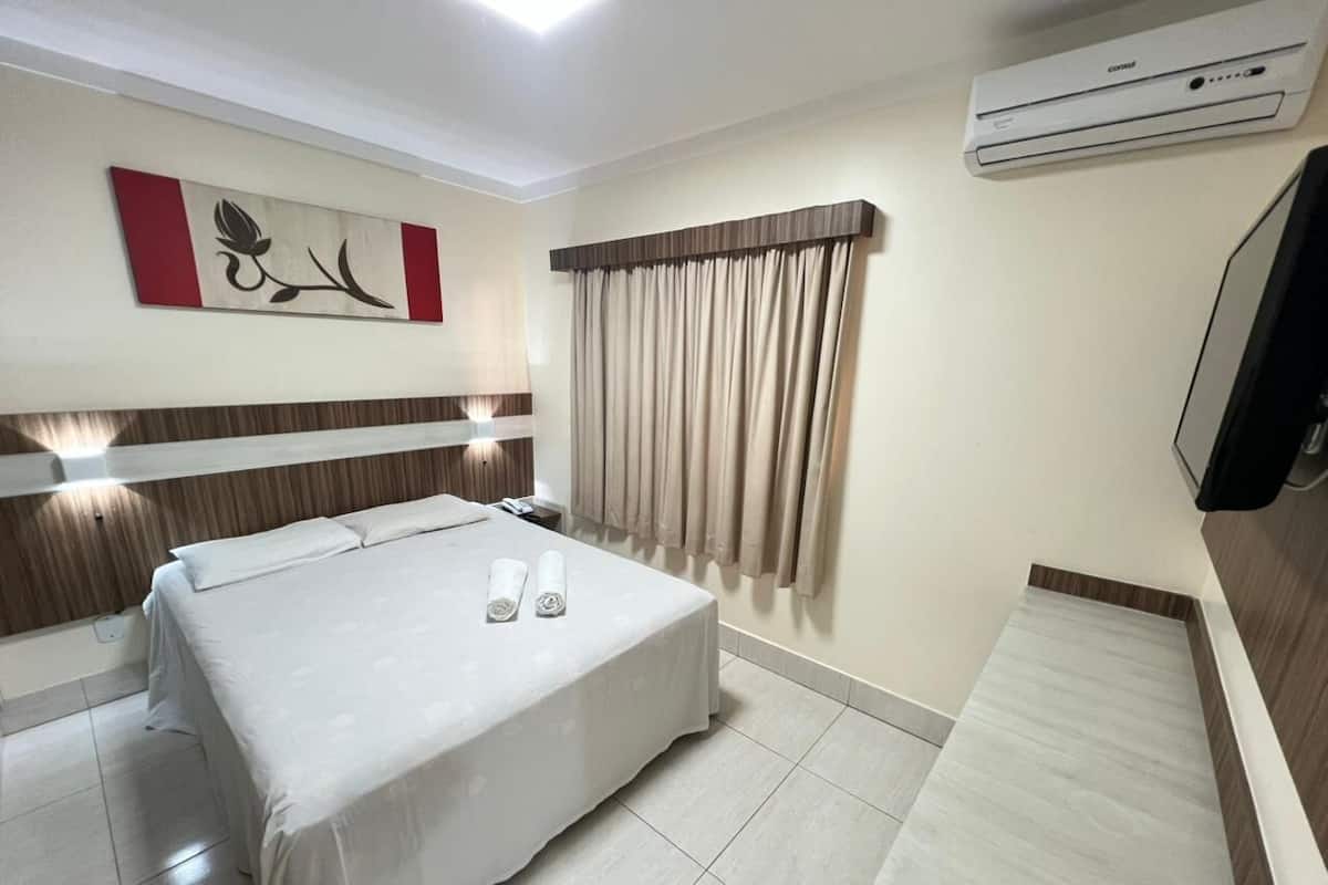 Ein Hotelzimmer mit einem Bett, einem Nachttisch, einer Wand-Klimaanlage und einem großen Fenster mit Vorhängen.