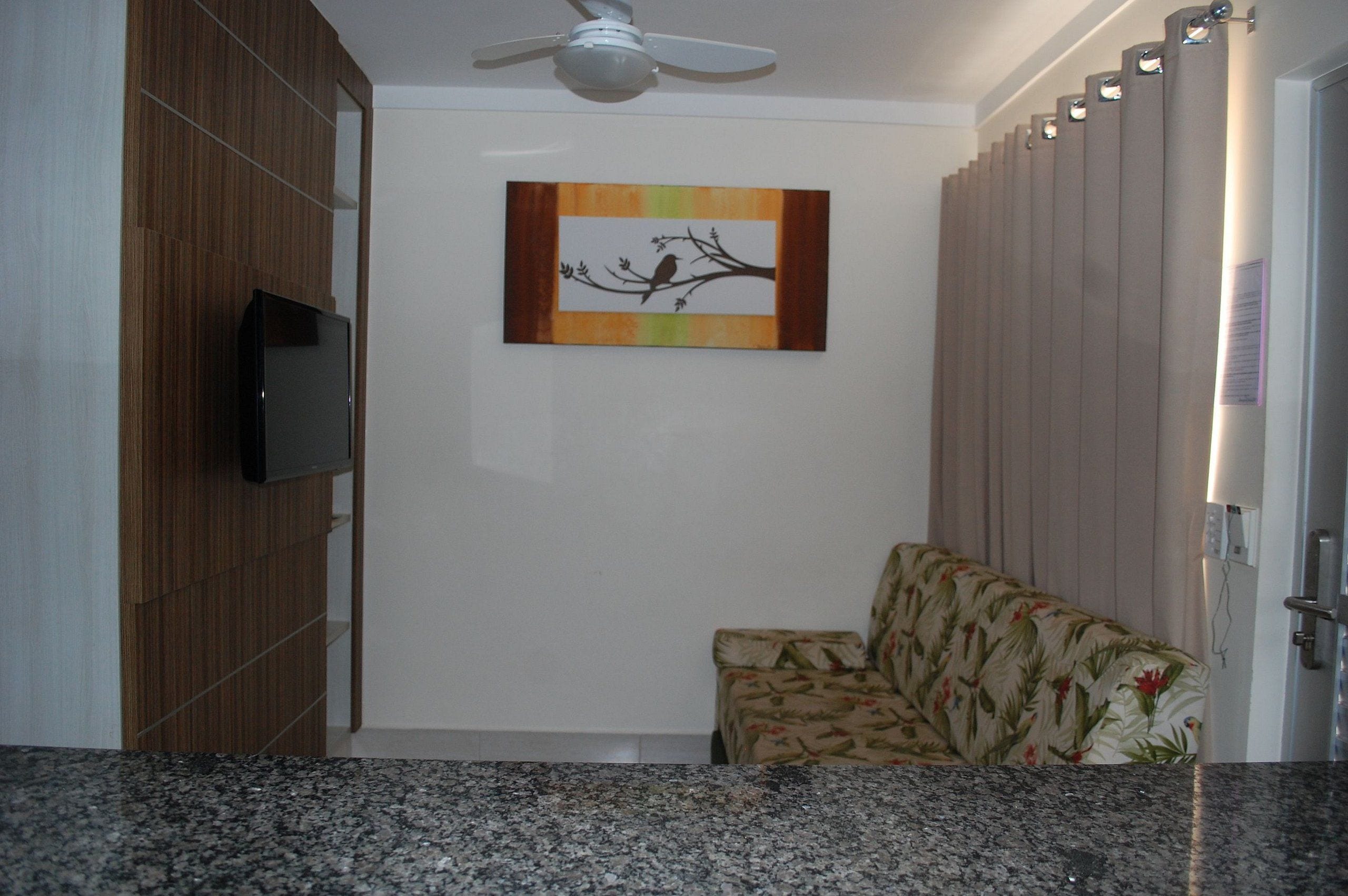 Ein Zimmer mit Deckenventilator, einem an einer Holzvertäfelung angebrachten Flachbildfernseher, einem sofaartigen Sitzmöbel mit Blumenmuster und einem an der Wand hängenden Kunstwerk mit einem Vogel auf einem Ast.