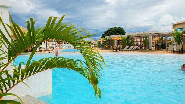 A swimming pool with lounge chairs and umbrellas, surrounded by palm trees and a building with a tiled roof. - Apartment Lacqua DiRoma IV - JC Temporada (Caldas Novas)