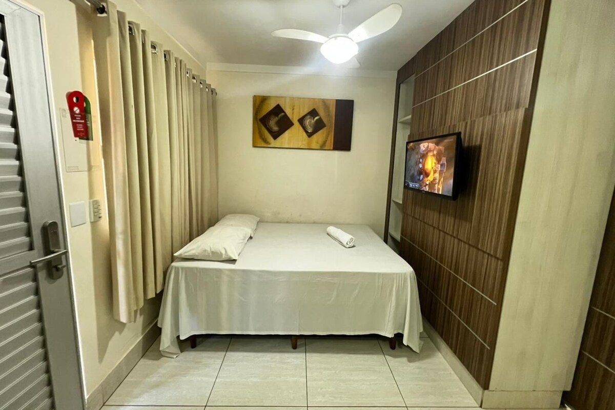 A hotel room with a bed, curtains, a ceiling fan, a flat-screen TV, and a wooden panel wall.