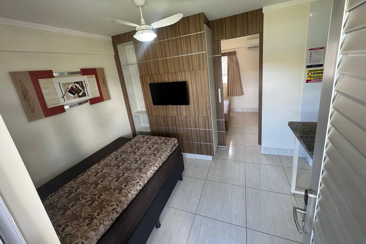 Een slaapkamer met een bed, een plafondventilator, een televisie en een badkamer met douche en toilet.