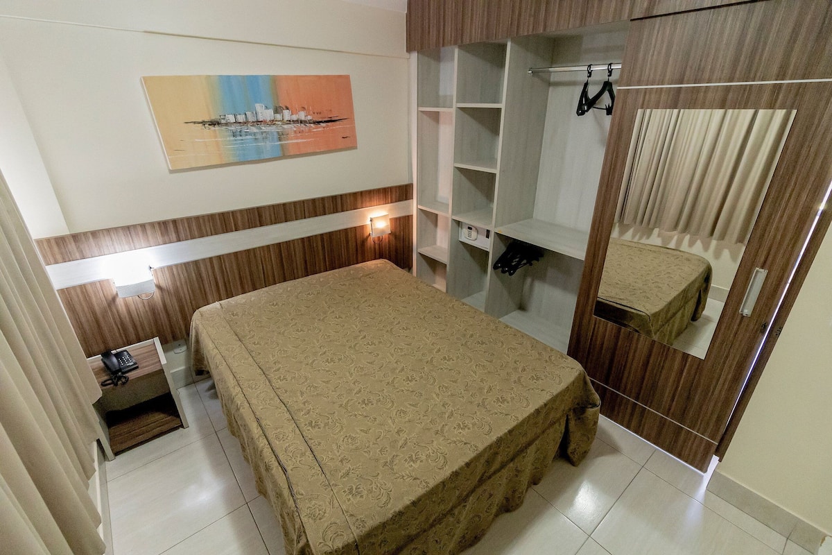 Een hotelkamer met een bed, een klein bureau met telefoon, een garderobe met spiegel en een schilderij aan de muur.