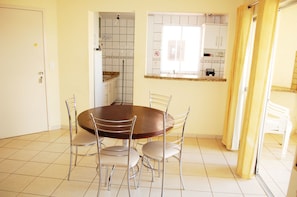 A dining area with a round table and four chairs, a kitchen with white cabinets, and a window with a curtain.