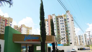A hotel named 'Vitoria DAS THERMAS' with a green facade, tall cypress trees, and a parking area with a few cars.