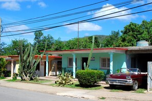 A vintage red car parked in front of a two-story building with a red roof and a small garden with cacti and a bush.