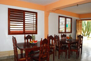 A dining area with a wooden table and chairs, a fruit bowl, and two windows with wooden blinds.