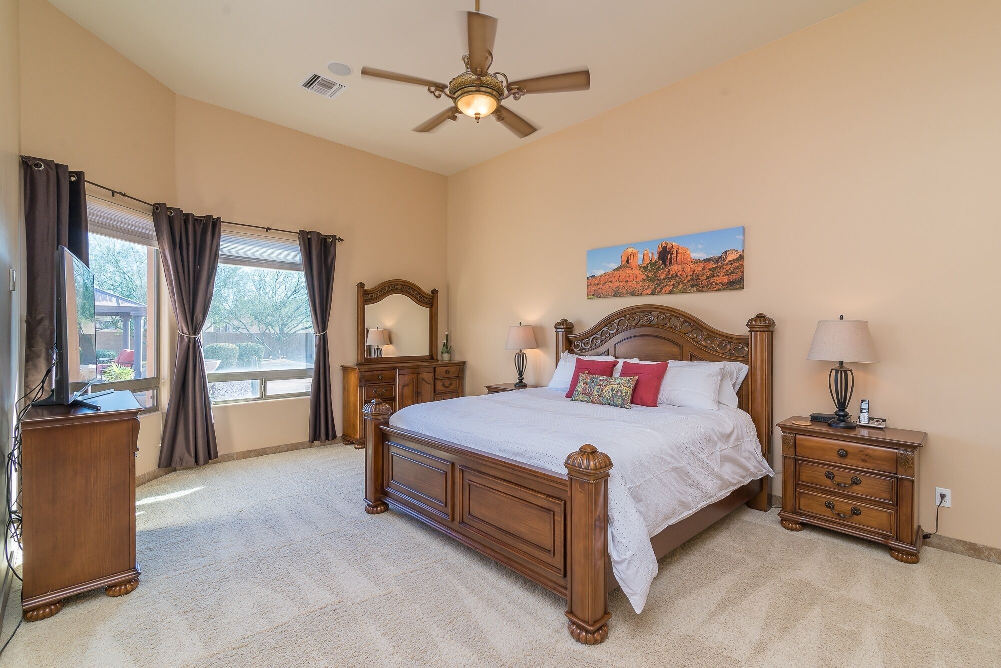 A bedroom with a large bed, a ceiling fan, a nightstand with a lamp, a dresser, and a window with curtains.