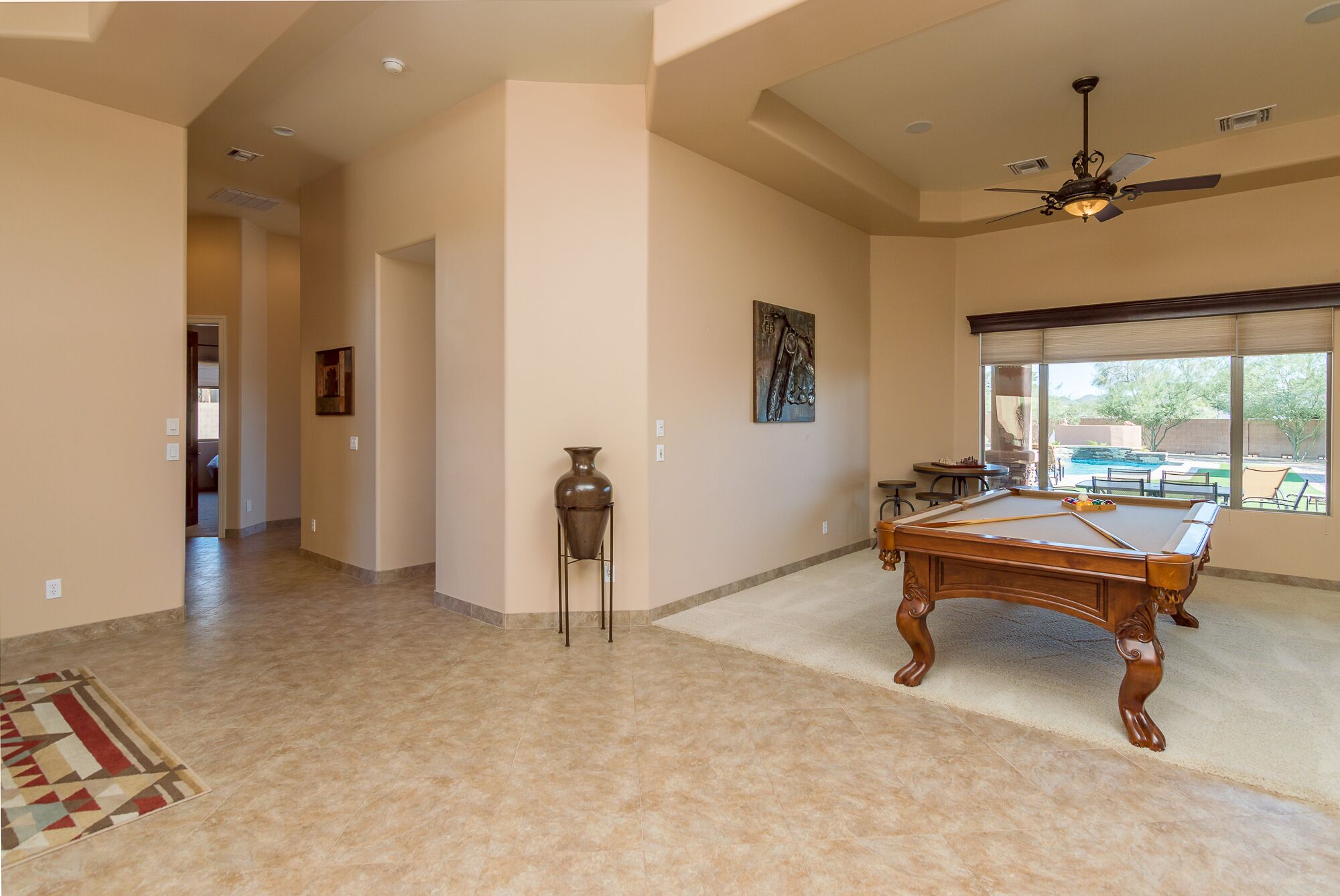 A spacious living area with a pool table, a large window with a view of a pool and outdoor seating, and a ceiling fan.