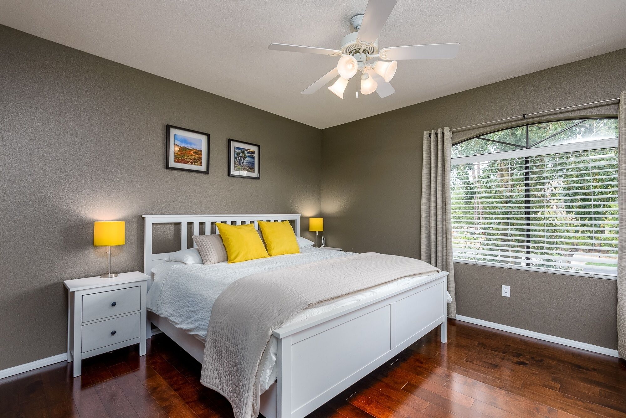 A bedroom with a white bed, two bedside lamps, a ceiling fan, and a window with blinds.