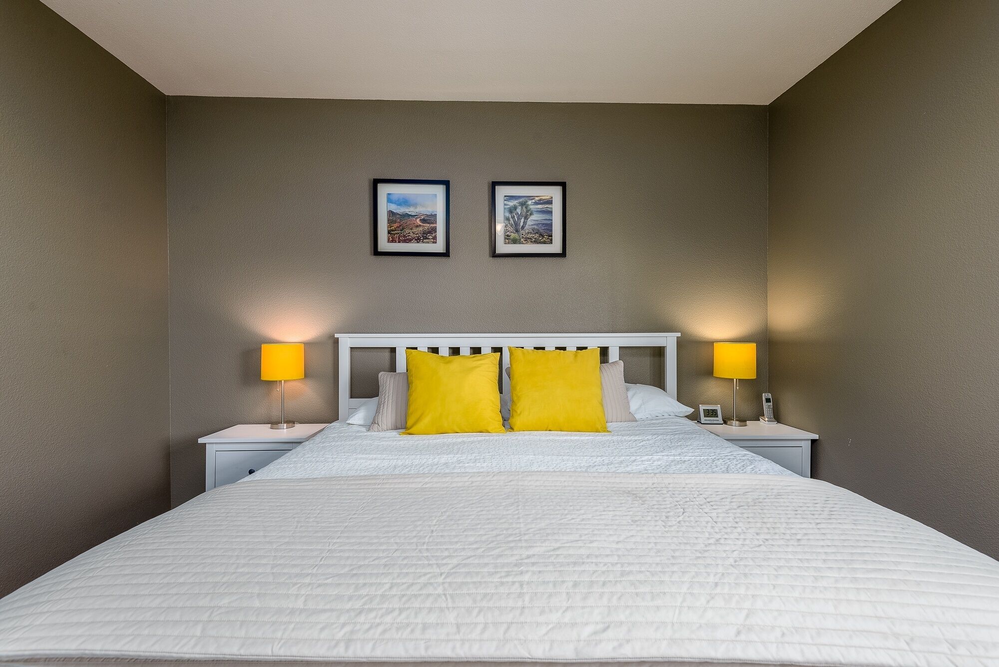 A neatly made bed with a white headboard, two bedside lamps, and two framed pictures on the wall.