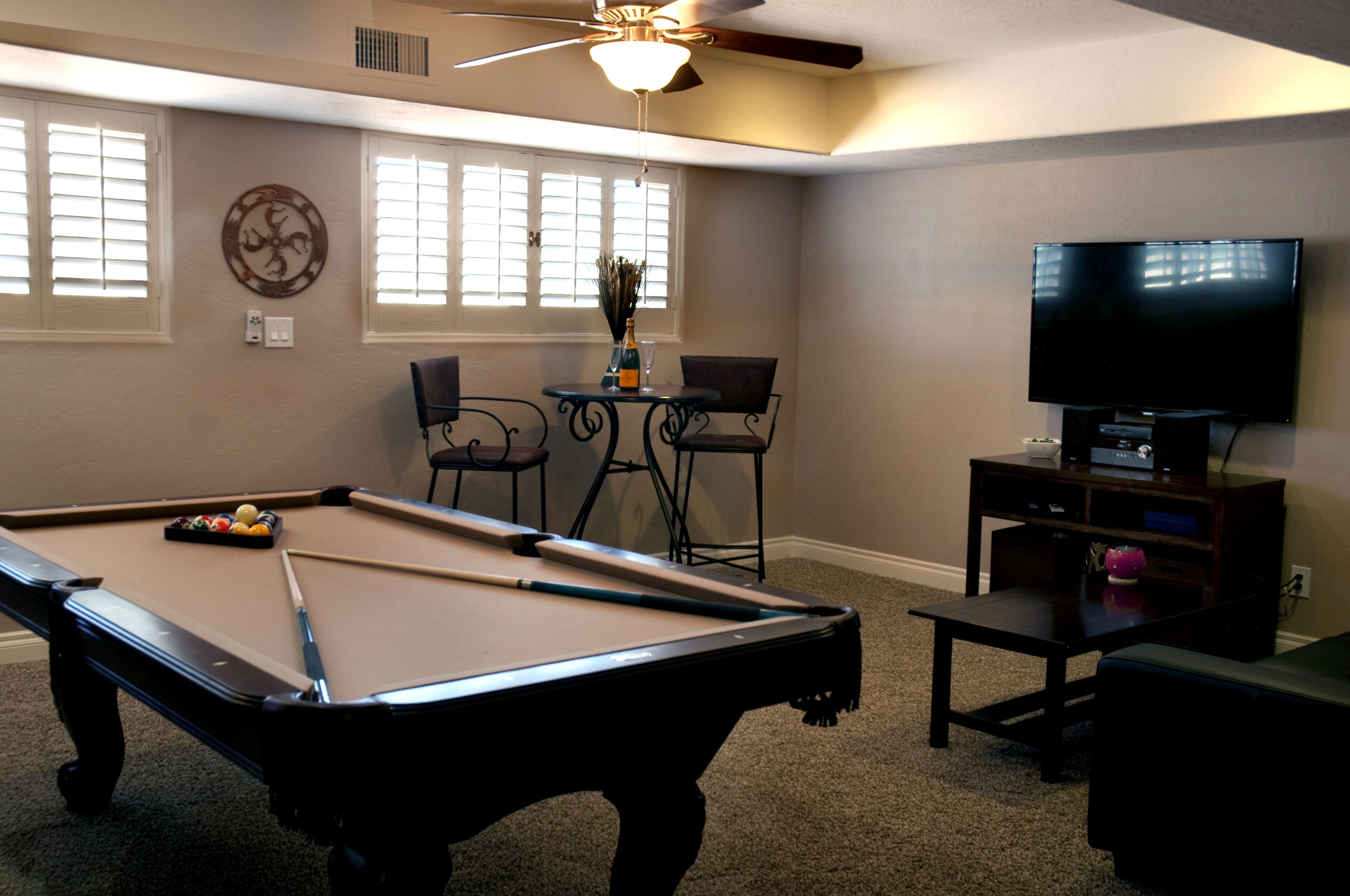 A pool table with balls, a television, and a ceiling fan in a room with windows and a small table with chairs.