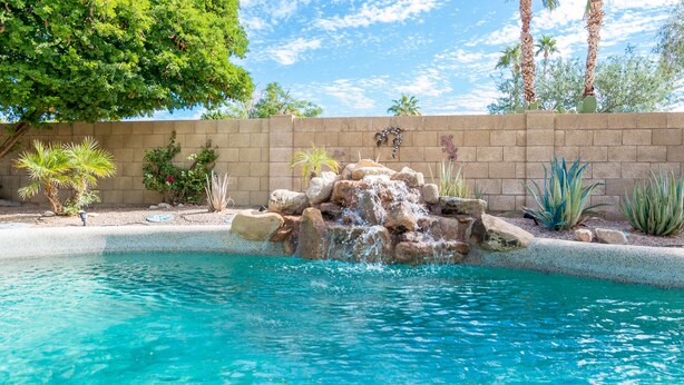 Une piscine agrémentée d’une cascade artificielle en roche, entourée d’un jardin aménagé avec différentes espèces de plantes et bordée d’un mur en briques.