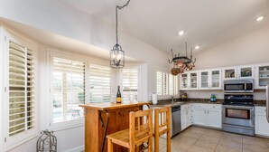 Une cuisine avec un bar en bois, des meubles blancs, des appareils électroménagers en acier inoxydable et de grandes fenêtres équipées de volets.