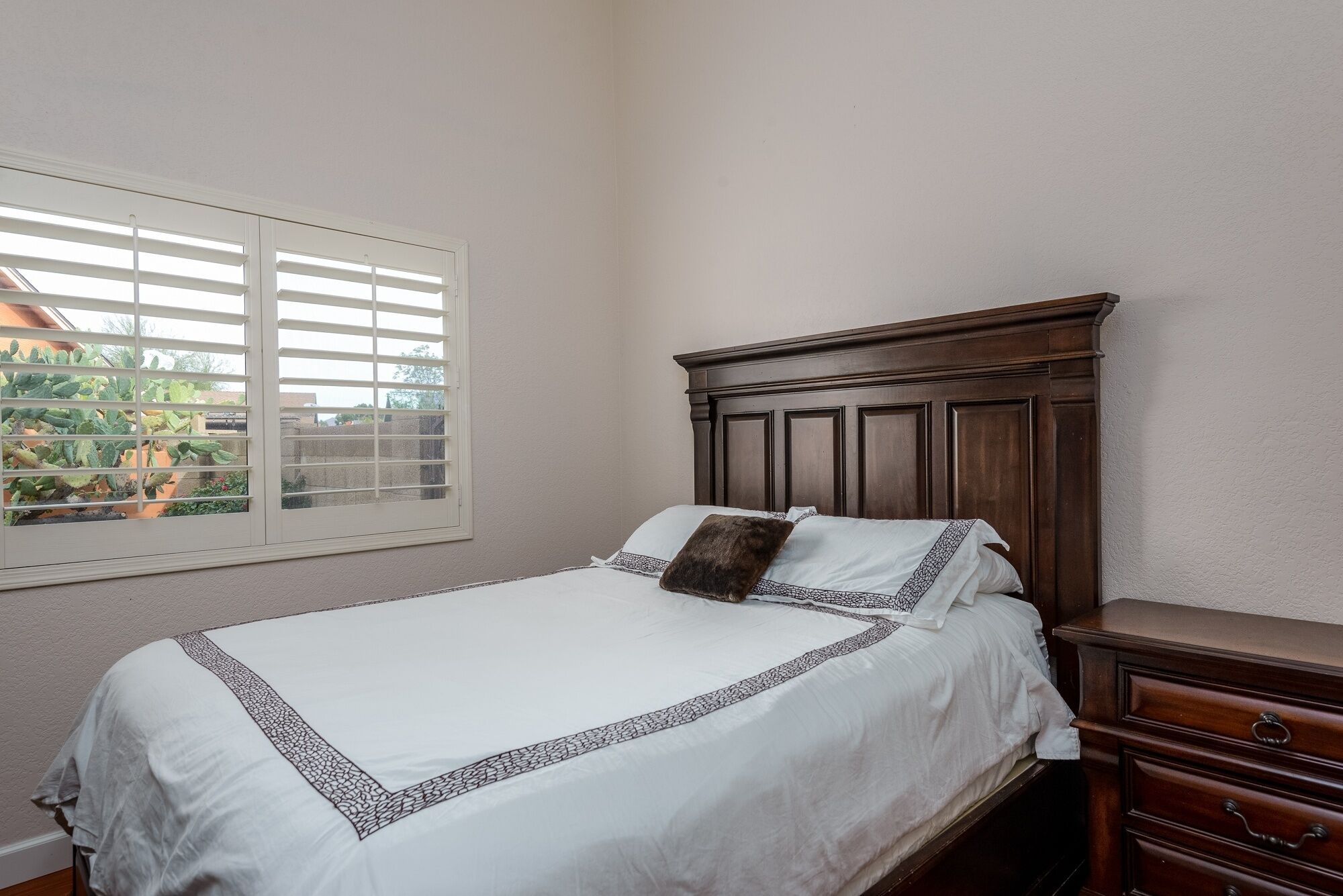 Une chambre à coucher avec une tête de lit en bois, un couvre-lit blanc orné d’une bordure décorative, un seul oreiller marron et une fenêtre avec des stores blancs.