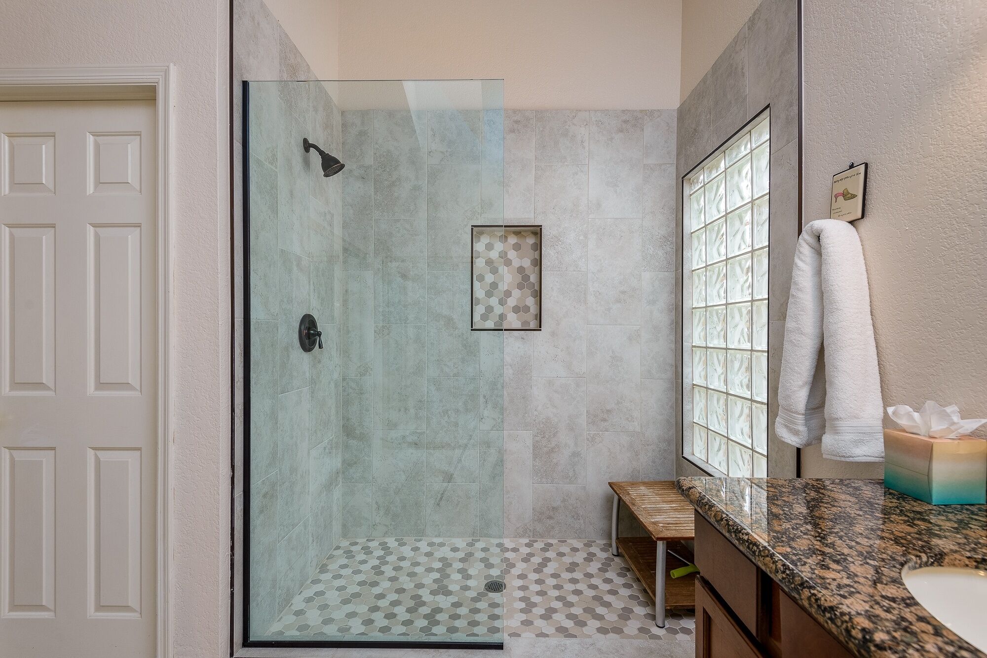 A bathroom with a glass shower enclosure, a wooden bench, a sink with a granite countertop, and a towel rack.