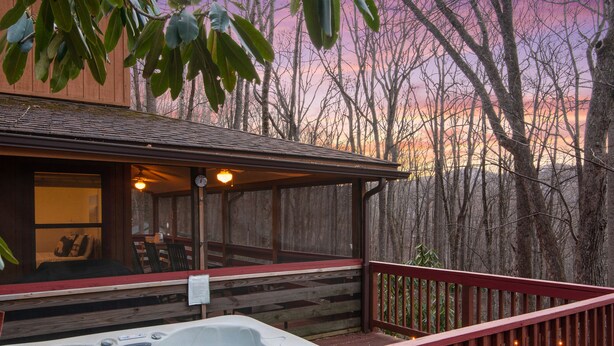 A wooden deck with a hot tub, a covered seating area, and a view of trees and a sunset. - Hot Tub - Screen Porch - Rockers - Fire Pit - Cozy - Clean - Fast Wi-Fi - 5 Star (Maggie Valley)