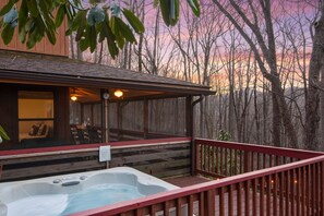 Eine Holzterrasse mit Whirlpool, überdachter Sitzgelegenheit und Blick auf Bäume und Sonnenuntergang.