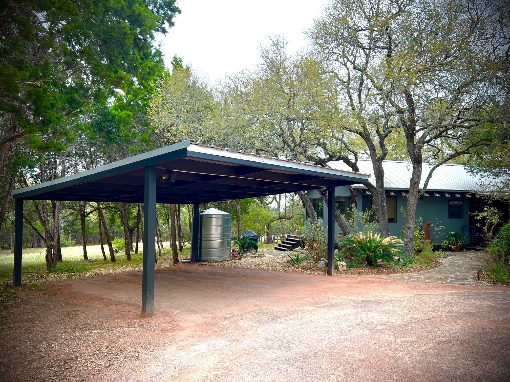A covered outdoor area with a metal water tank, surrounded by trees and a house in the background.