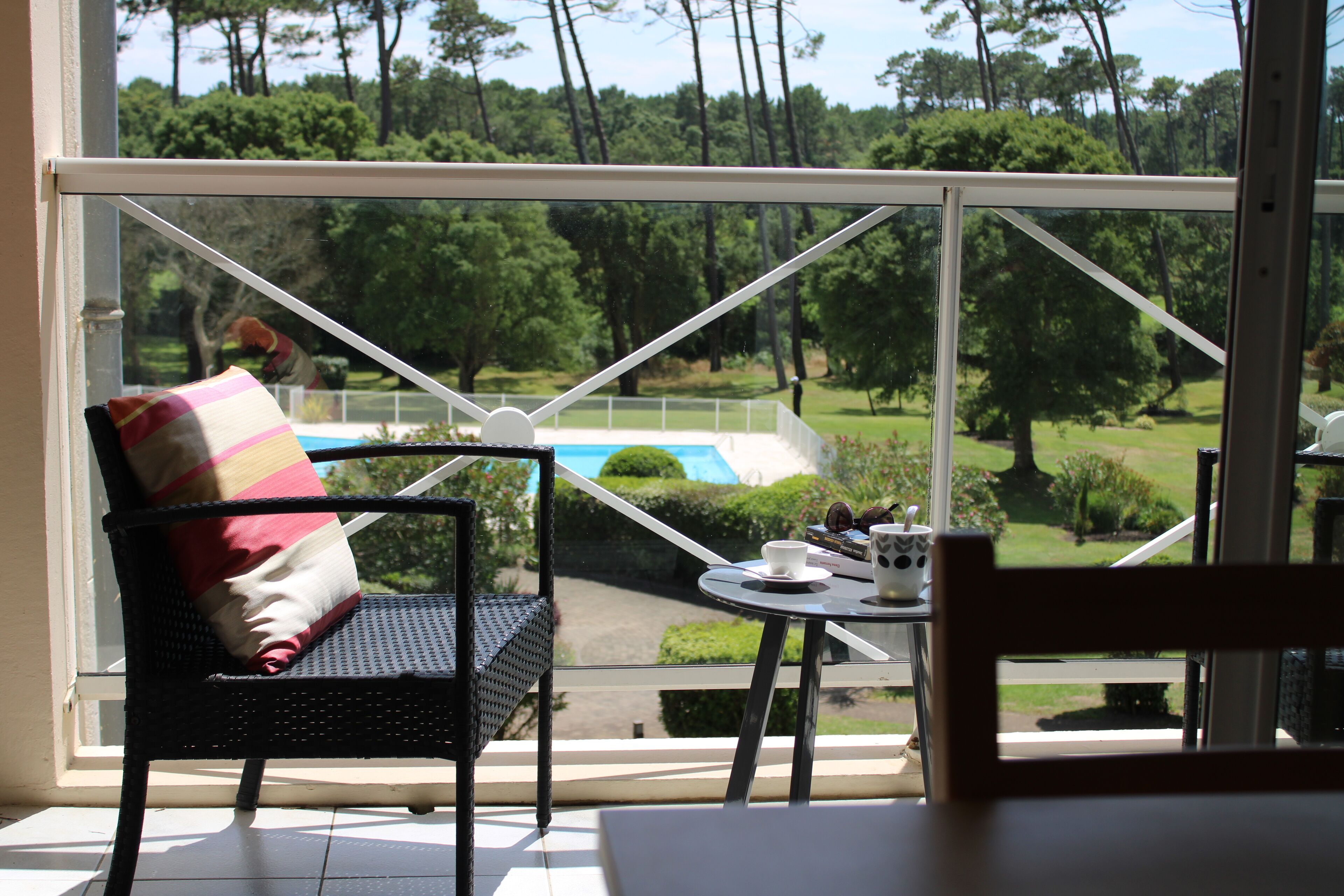 A room with a view of a pool and greenery, a wicker chair with a cushion, and a small table with a cup and saucer.