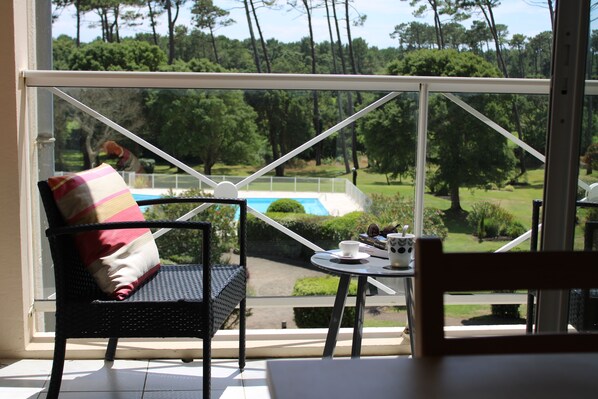 Habitación con vistas a una piscina y vegetación, una silla de mimbre con cojín y una mesita con taza y platillo.