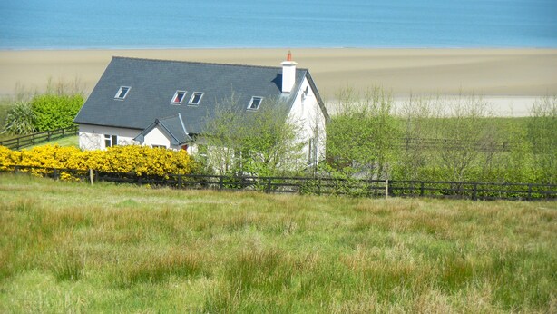 A house with a sloped roof, a chimney, and a small porch. It is surrounded by a wooden fence and a hedge of yellow flowers. - Stunning Beachfront Cottage  Ballybunion (Ballybunion,)
