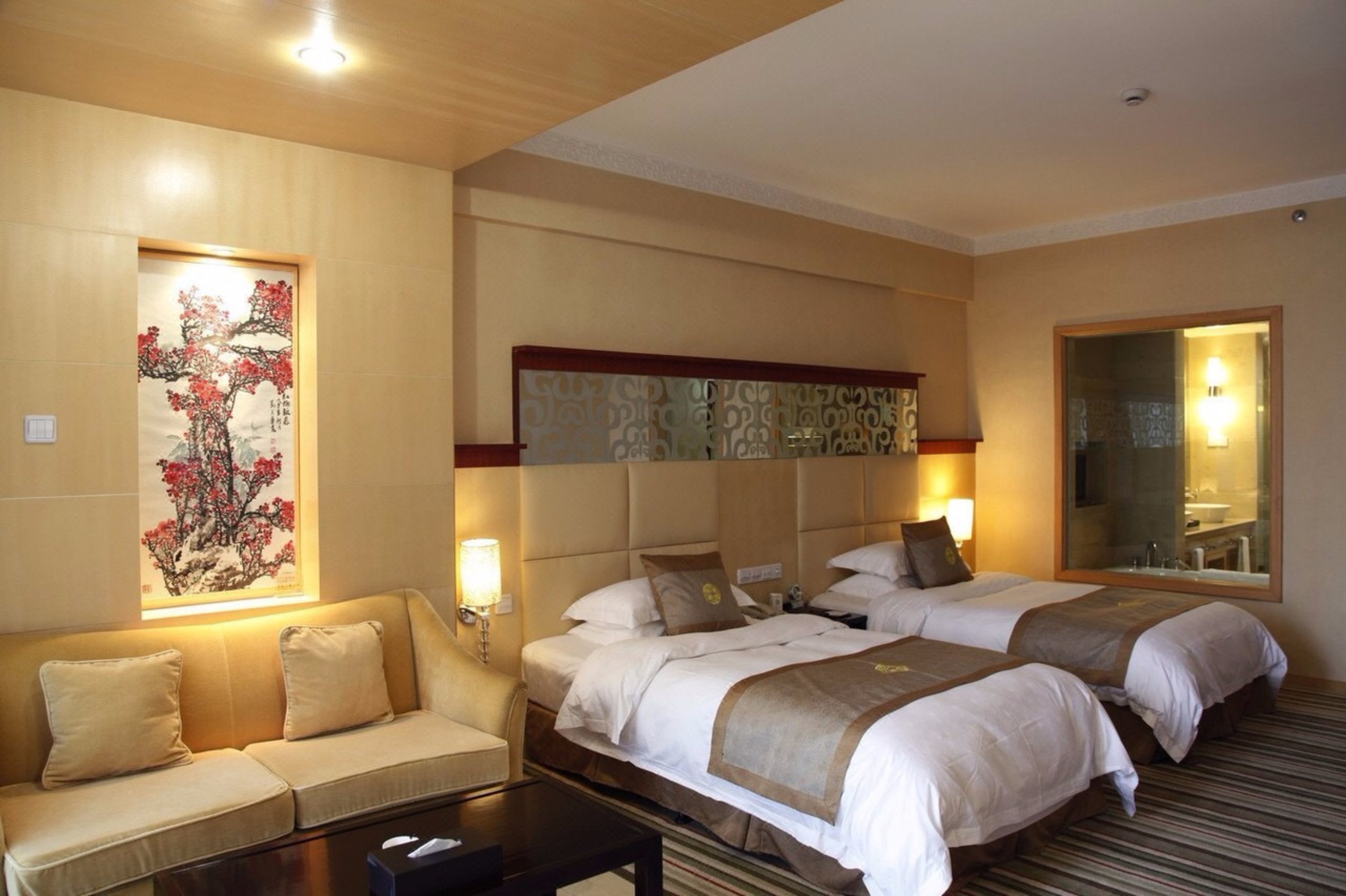 A hotel room with two beds, a sofa, a large artwork on the wall, and a mirror reflecting the bathroom.