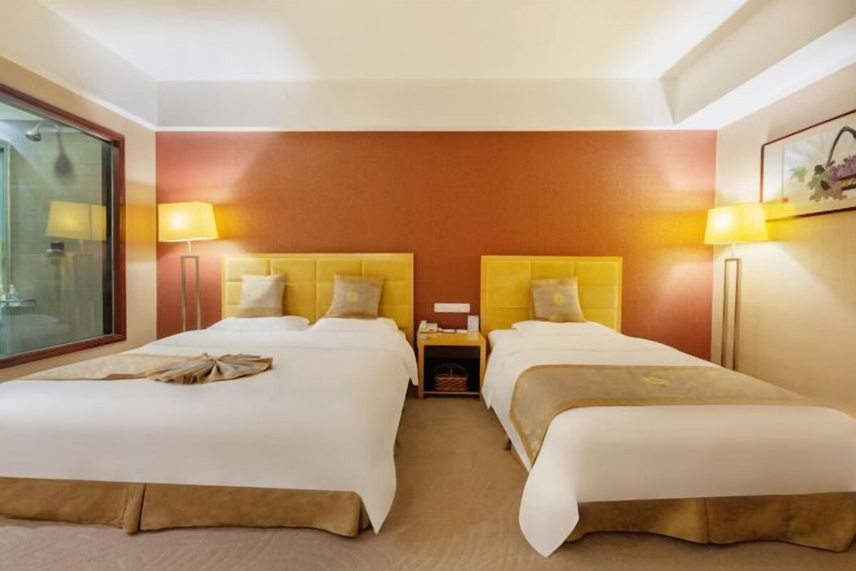 A hotel room with two beds, each with a headboard and bedside lamps. There is a small table between the beds and a window on the left side.