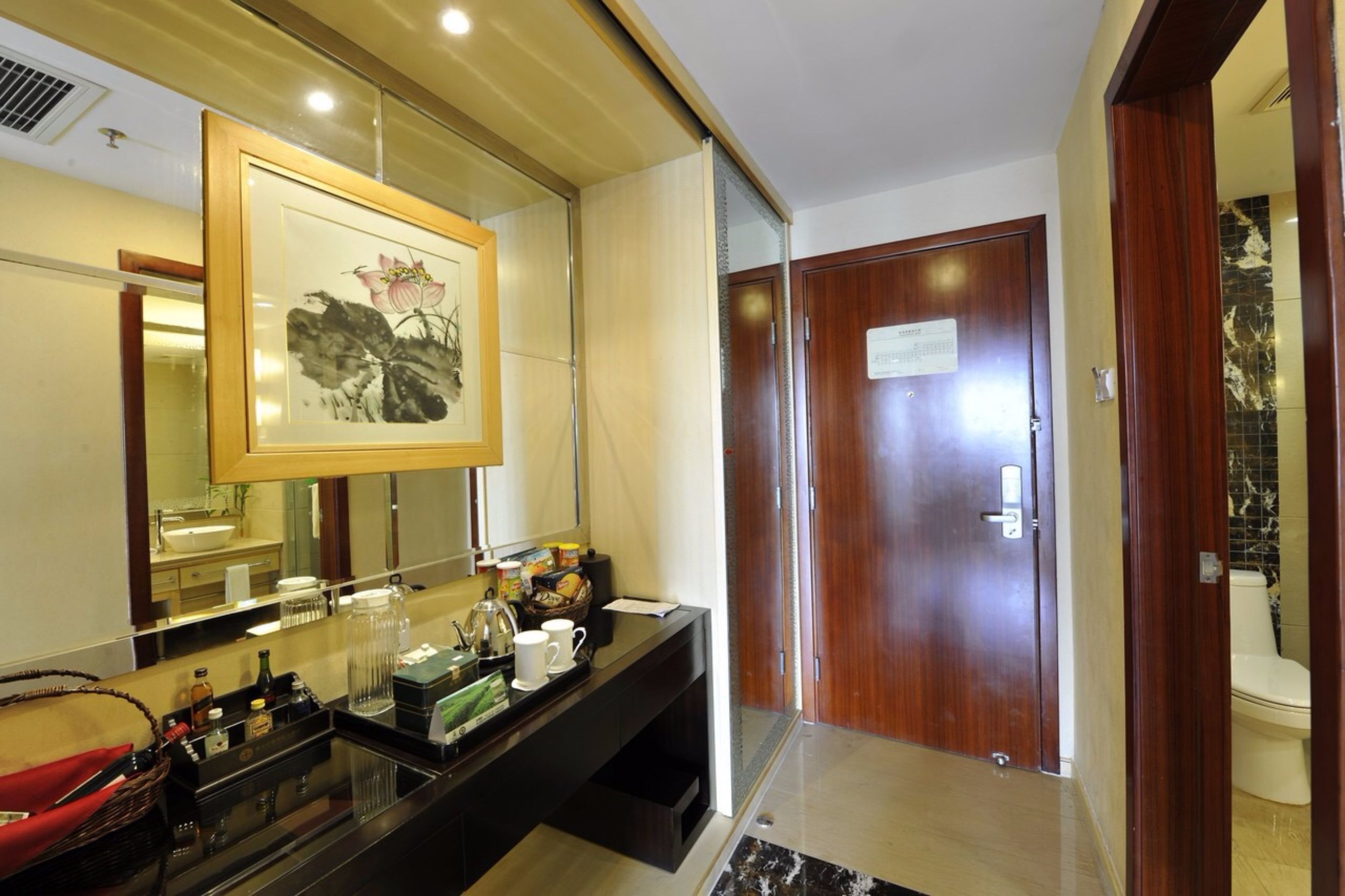 Une salle de bain d’hôtel avec un grand miroir, une œuvre d’art encadrée, une commode avec des articles de toilette et une porte en bois.