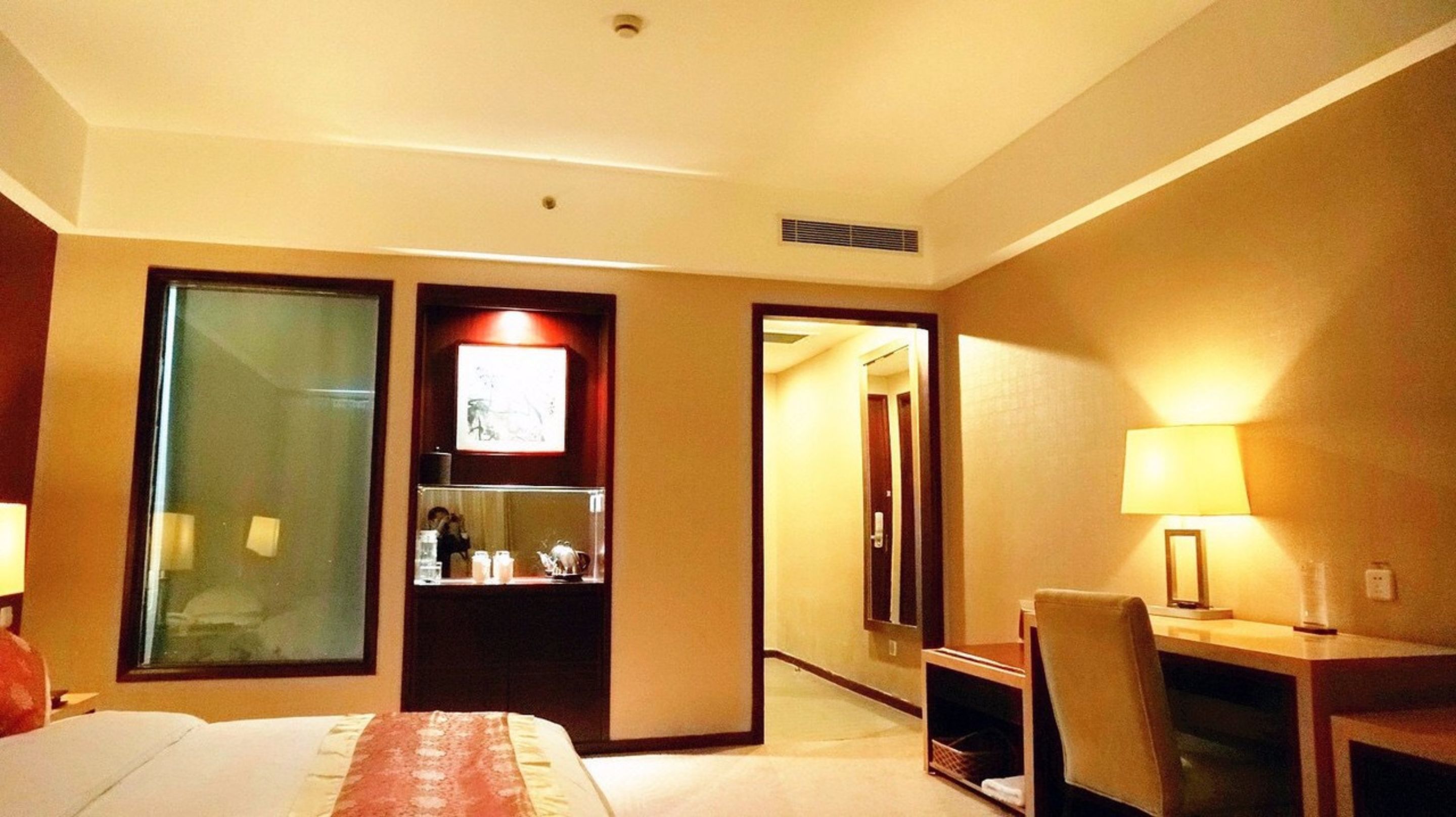 A hotel room with a bed, a desk, a chair, a desk lamp, a mirror, and a door leading to another room.