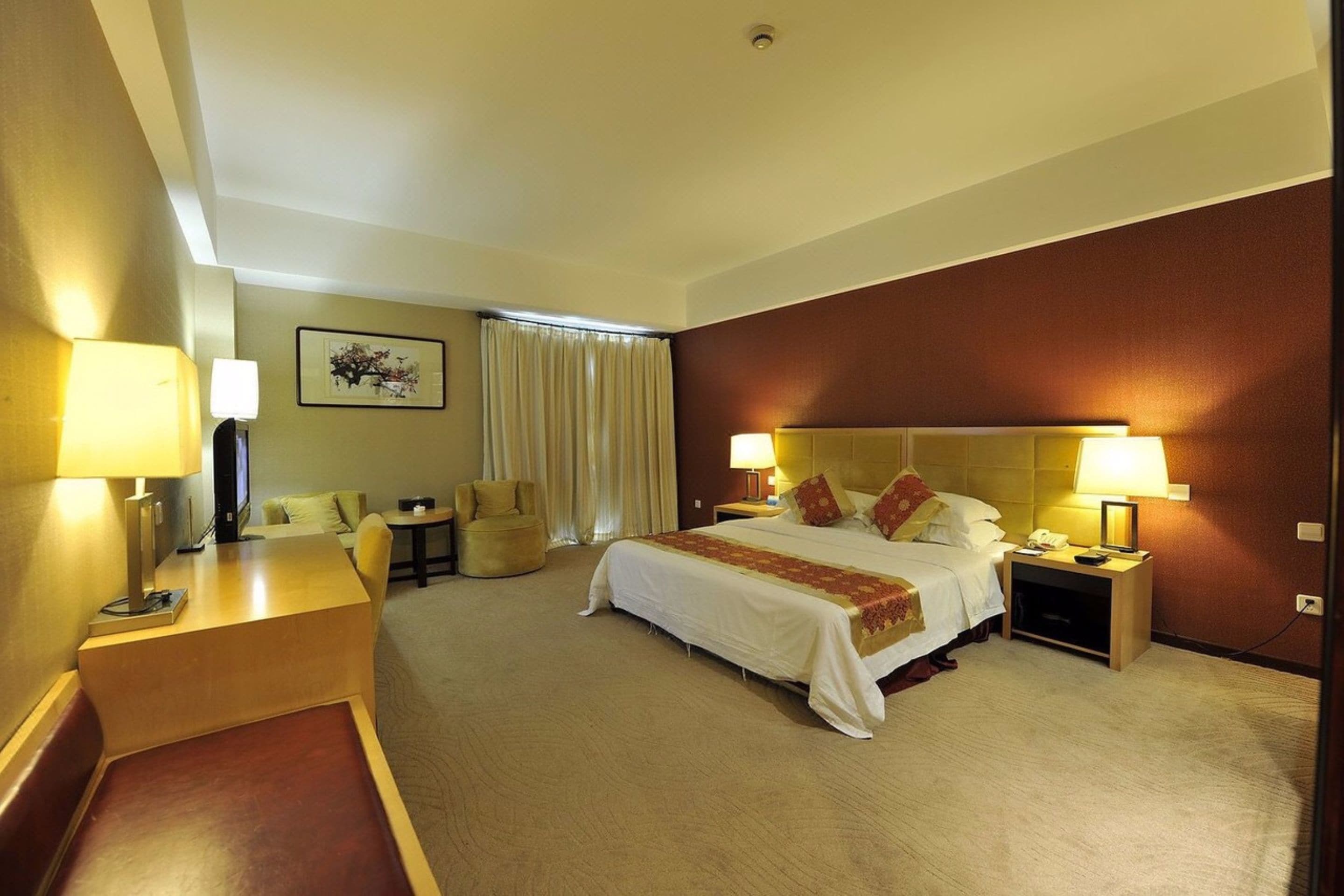 A hotel room with a large bed, two bedside tables with lamps, a desk with a chair, a small table, and a large window with curtains.