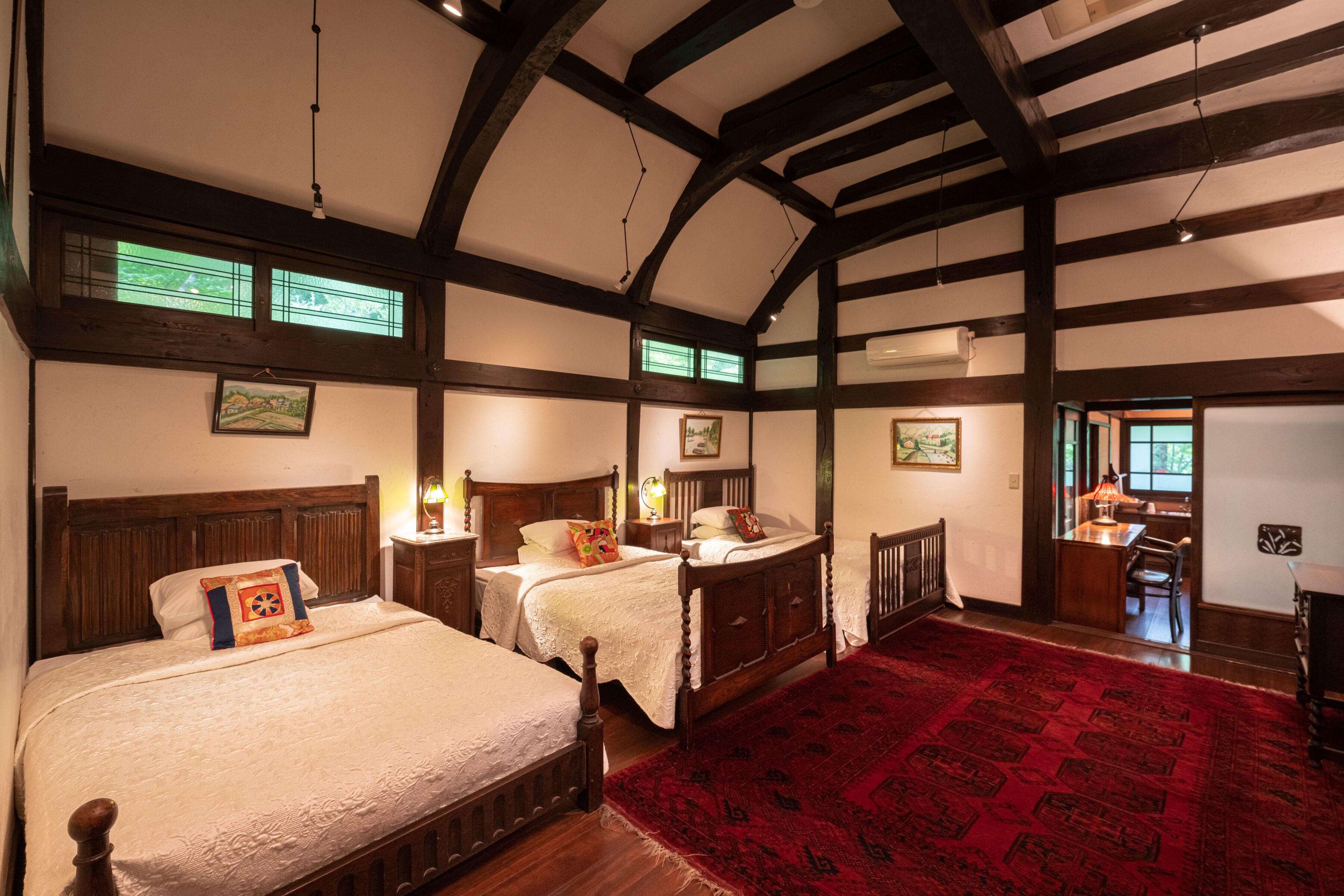 A traditional room with two wooden beds, a red carpet, and a view of a dining area through a window.