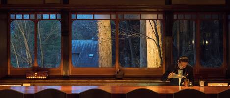Une pièce faiblement éclairée, avec une longue table et des chaises, de grandes fenêtres donnant sur des arbres et un bâtiment à l’extérieur.