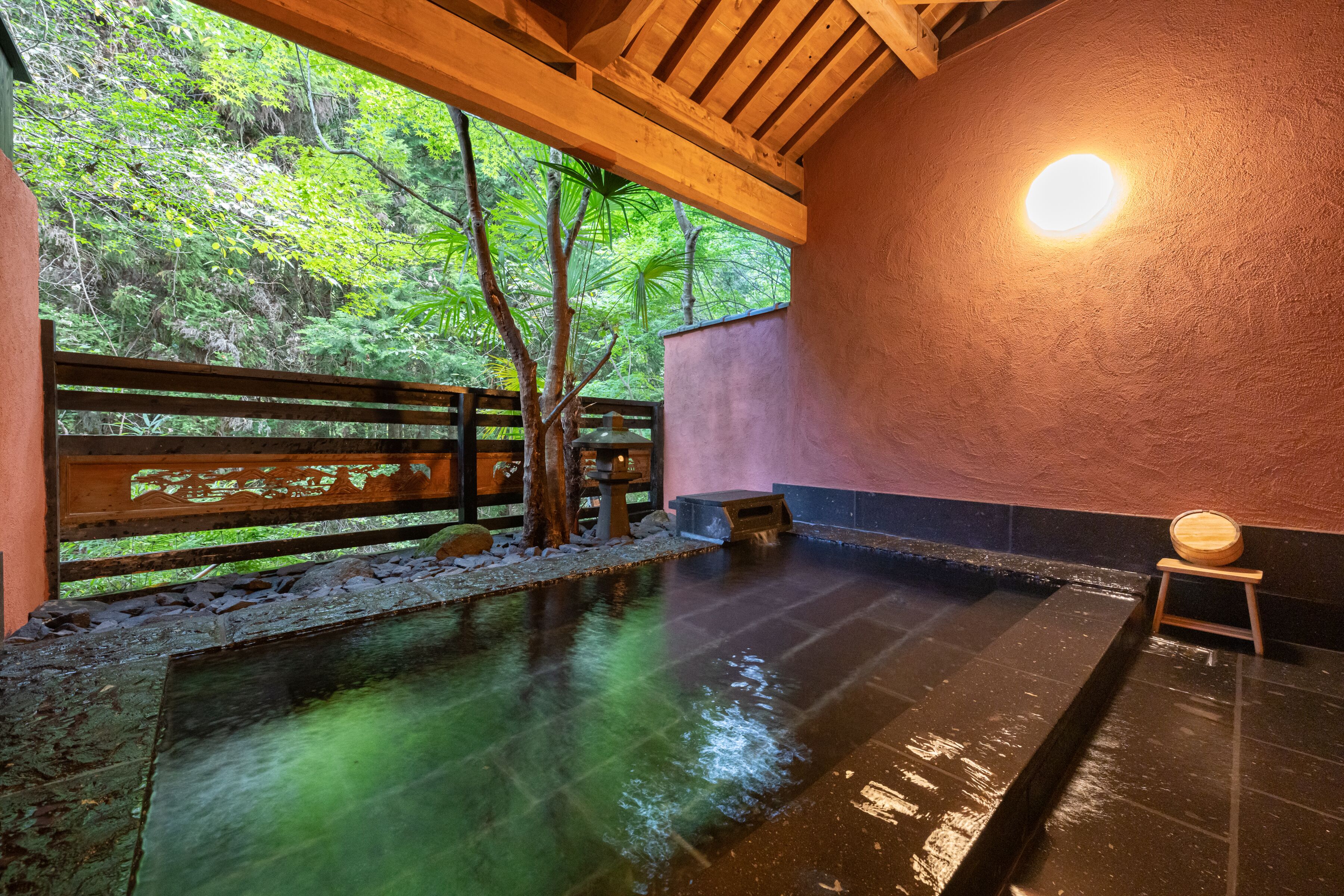 A traditional Japanese onsen with a wooden deck, a stone bath, and a view of lush greenery.