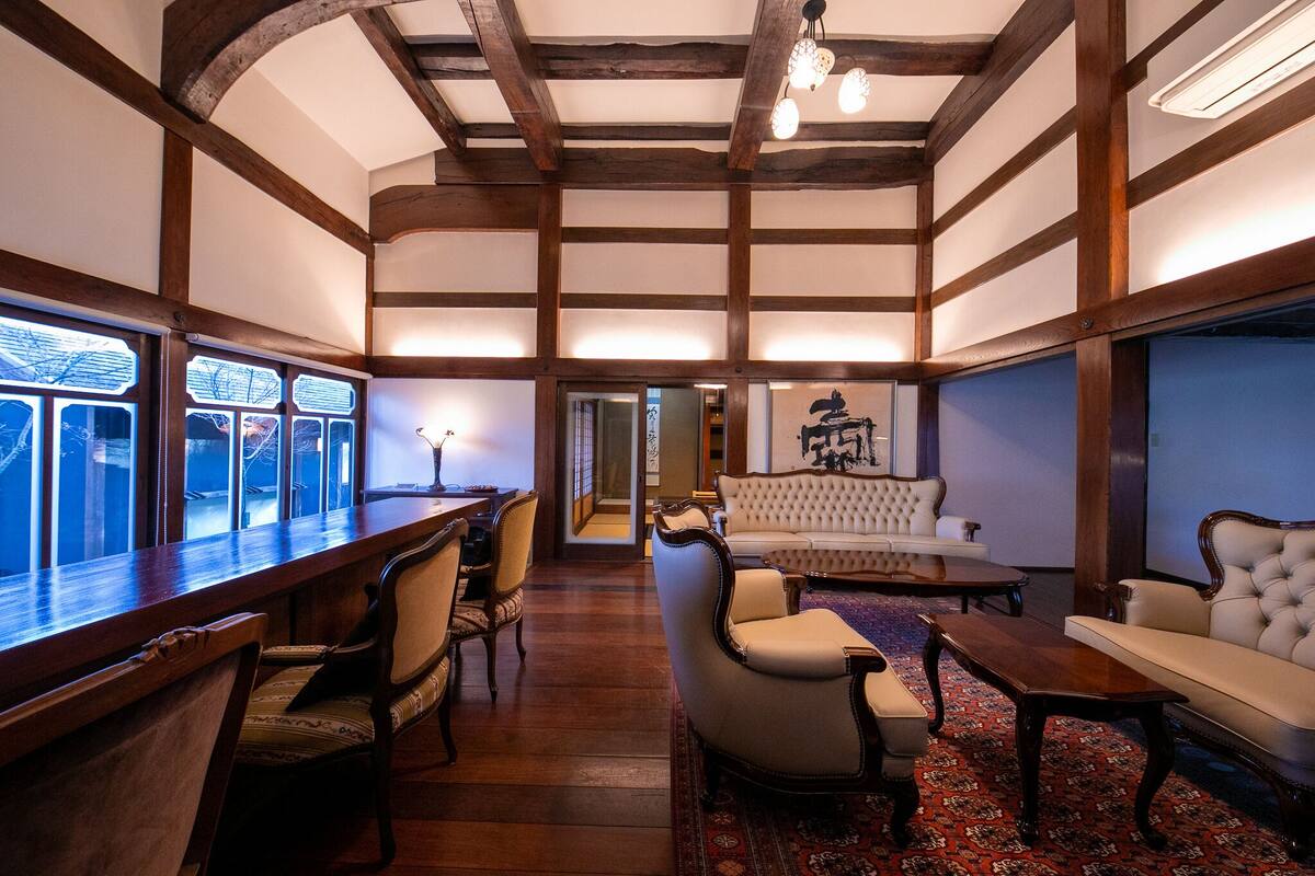 A traditional room with wooden beams, a sofa, chairs, a table, and a view through large windows.