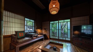 A traditional Japanese-style room with tatami flooring, a low wooden table with a kettle, a sofa with cushions, a television, and large windows with a view of greenery. - Nihon No Ashitaba (Yufu)