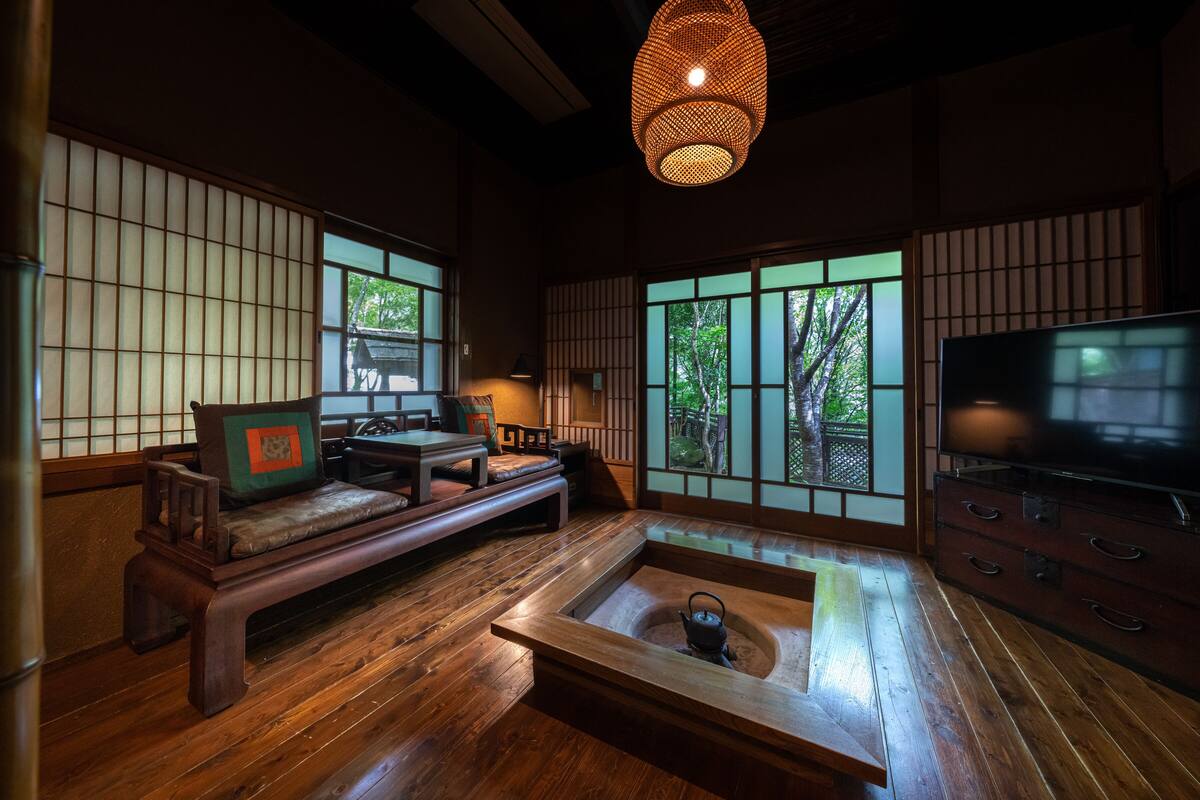 A traditional Japanese-style room with tatami flooring, a low wooden table with a kettle, a sofa with cushions, a television, and large windows with a view of greenery.