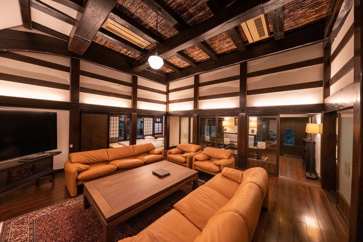A spacious living room with a large sectional sofa, a wooden coffee table, and a TV. The room features exposed wooden beams and a traditional Japanese aesthetic.