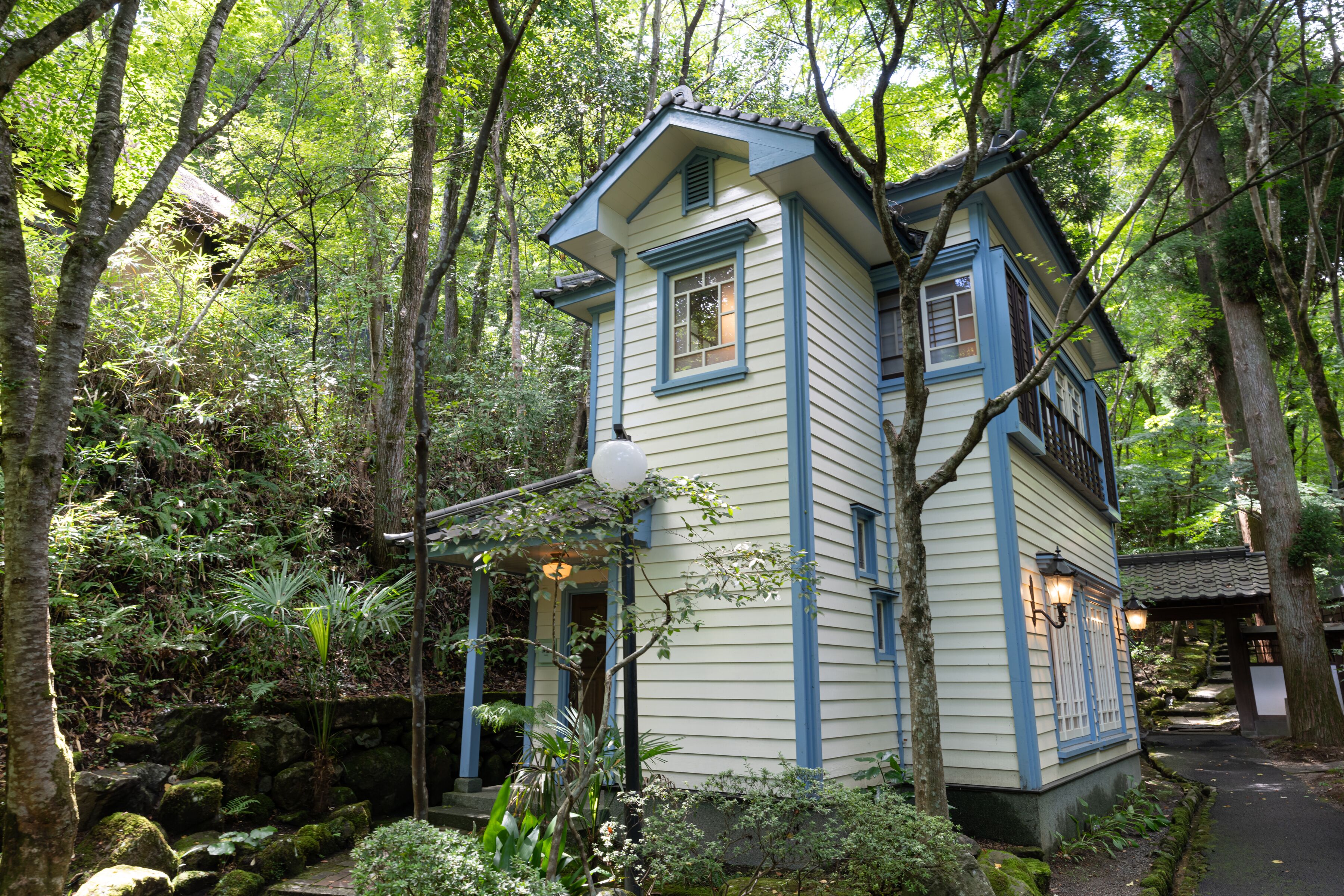 A small, two-story house with a steeply pitched roof, surrounded by dense forest.