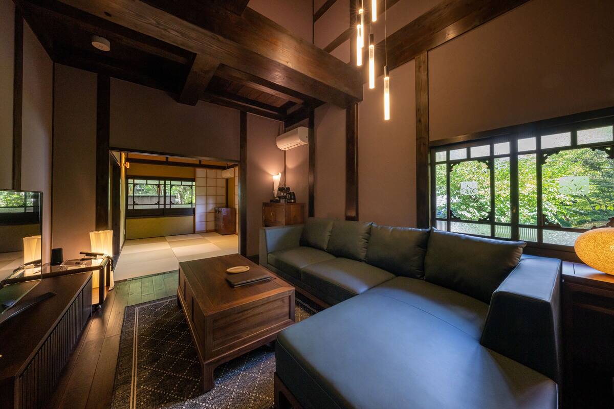 A modern living room with a sofa, coffee table, and a view of greenery through large windows.