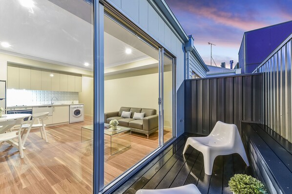 A modern apartment with a sliding glass door leading to a living area, a kitchen, and a balcony with outdoor seating. - Aloha Luxury Central Apartments (Mount Gambier)
