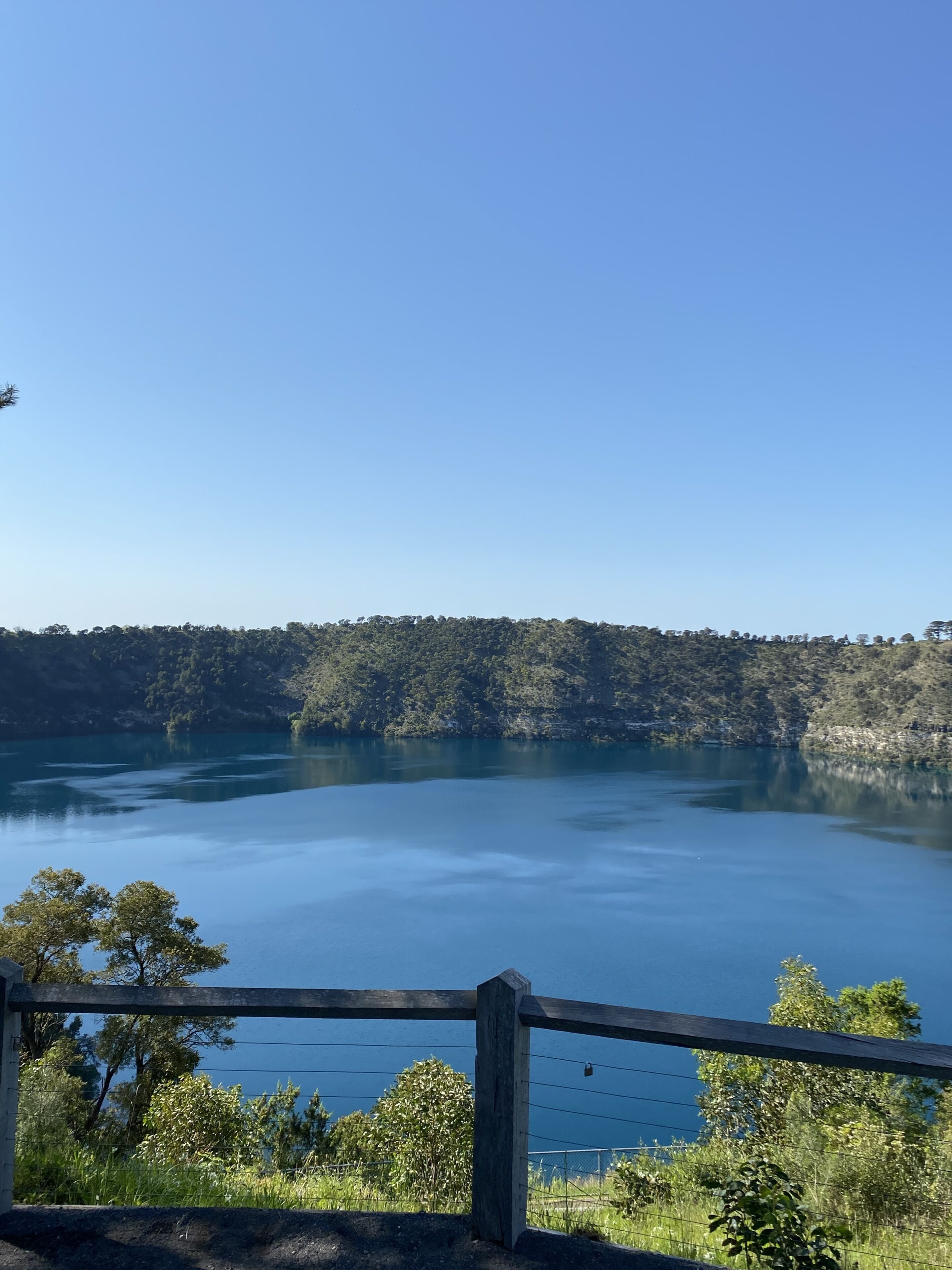 A serene lake with a wooden fence in the foreground, surrounded by lush greenery and hills in the background.